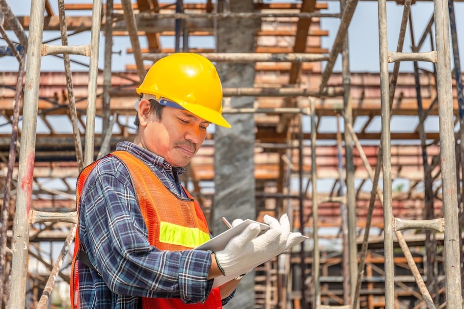 Construction worker wearing a hard hat reviews a clipboard checklist at a job site.